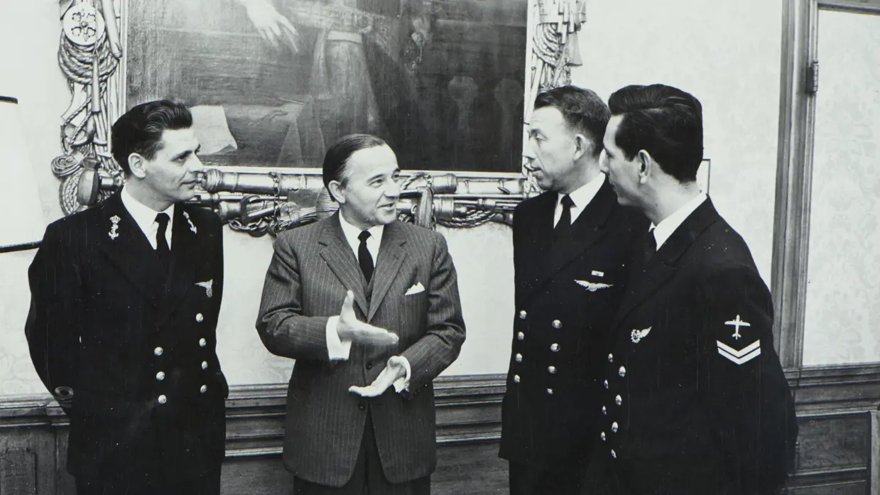 4 mannen in een formele, officieuze setting: 3 in donkere marine-uniformen met insignes, 1 in de middelste man draagt een pak. Op de achtergrond is een grote, ornamentele muurschildering zichtbaar.