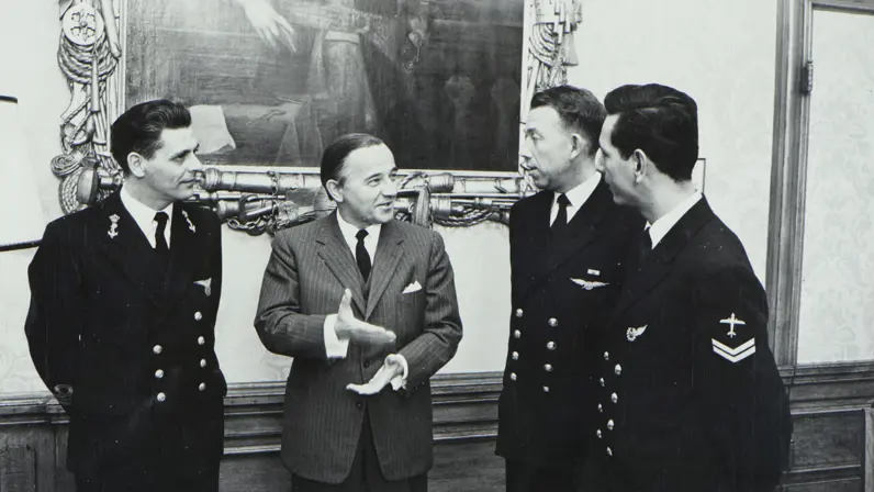 4 mannen in een formele, officieuze setting: 3 in donkere marine-uniformen met insignes, 1 in de middelste man draagt een pak. Op de achtergrond is een grote, ornamentele muurschildering zichtbaar.