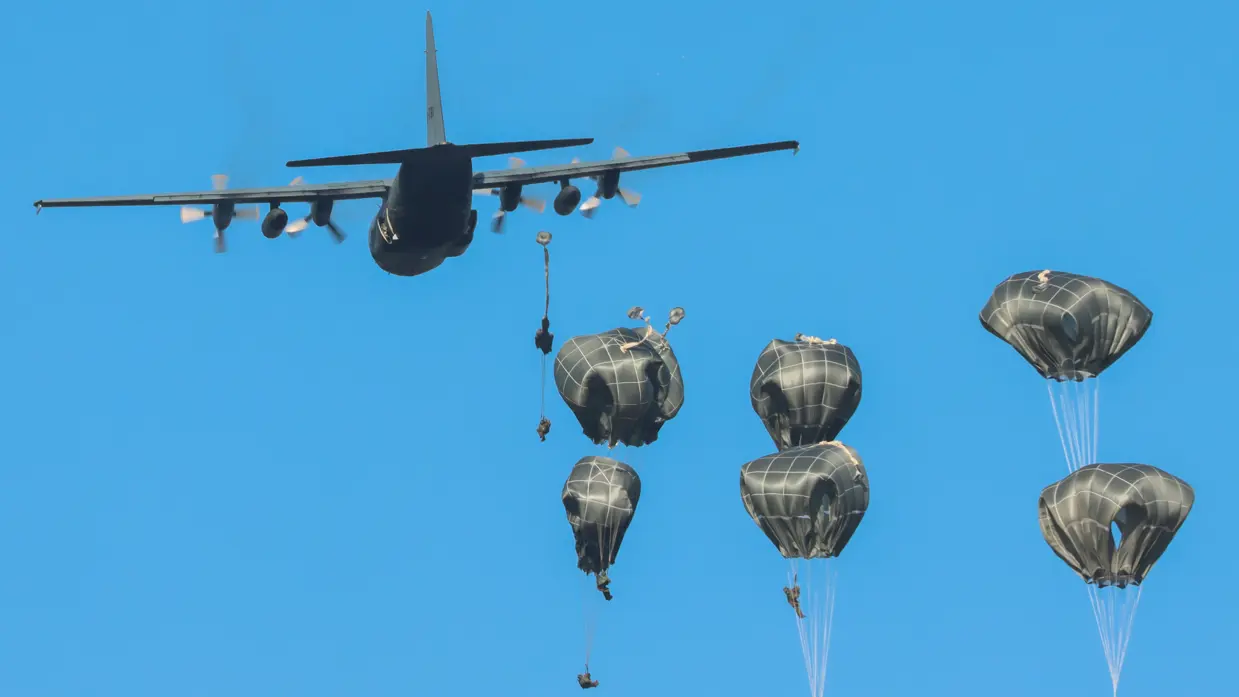 Gouden parachutes met militairen komen uit een militair vrachtvliegtuig onder een blauwe hemel.