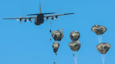 Gouden parachutes met militairen komen uit een militair vrachtvliegtuig onder een blauwe hemel.
