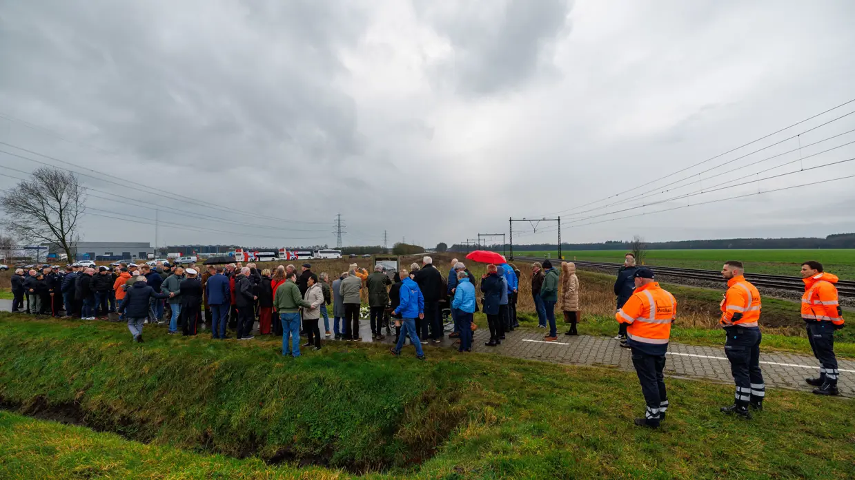 Links een groep mensen, waaronder militairen in uniform bij een bord langs de spoorlijn. Rechts: bloemen en kransen bij een bord met daarop een afbeelding van een oude trein, terwijl een moderne trein langs raast.