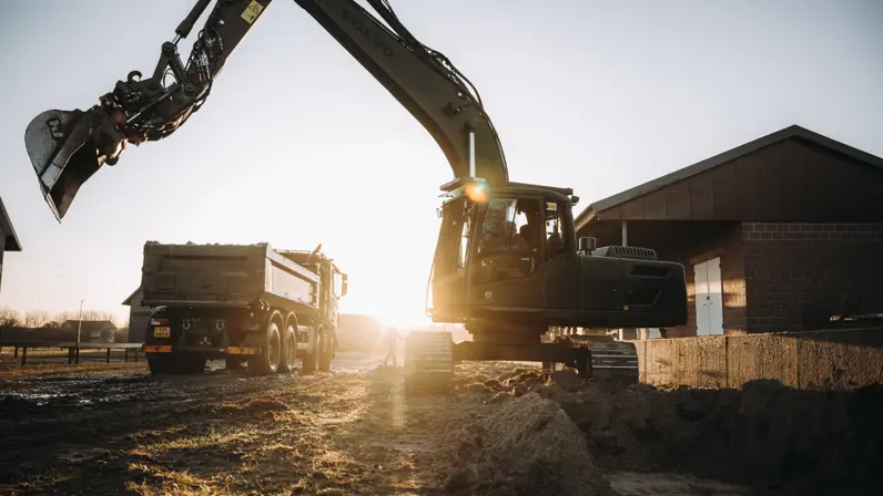  Een graafmachine verplaatst grond, terwijl een vrachtwagen dichtbij staat op een boerenerf, met de ondergaande zon in de achtergrond en gebouwen opzij.