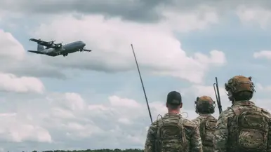 Drie militairen kijken vanaf de grond omhoog naar een vliegtuig dat overvliegt.