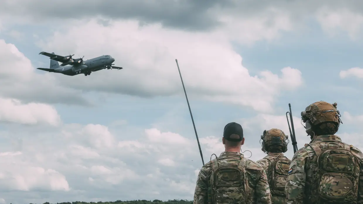 Drie militairen kijken vanaf de grond omhoog naar een vliegtuig dat overvliegt.
