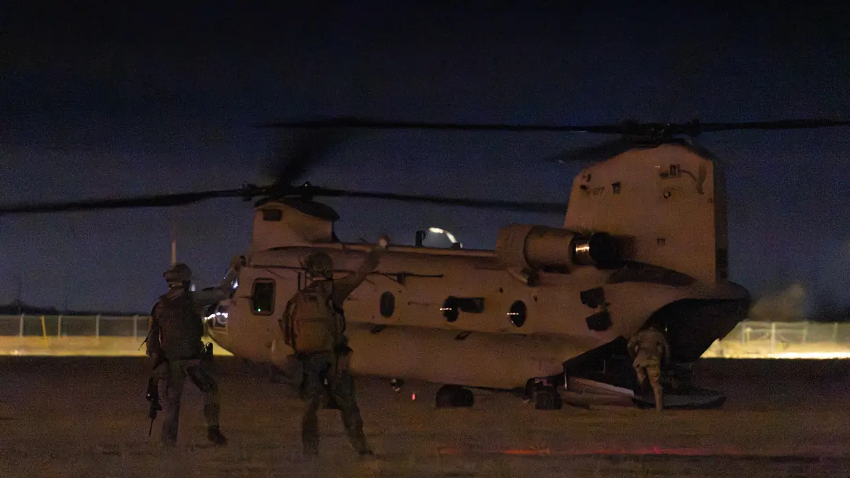 In schemering stapt laatste militair via de achterkant de heli in. Twee militairen op het terrein geven met gebalde vuist de vlieger een teken.