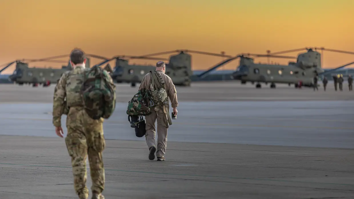 Twee militairen lopen met tassen richting drie Chinooks op een platform. Op de achtergrond een oranje-gele lucht.