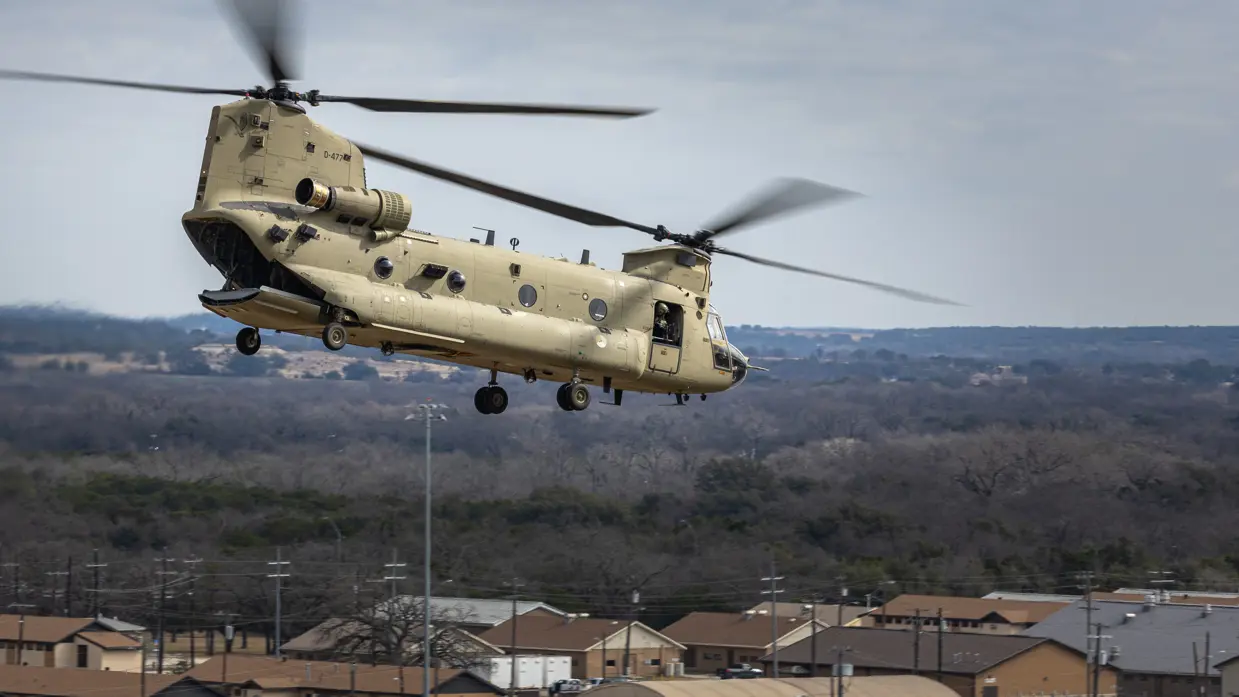 Laagvliegende Chinook over militaire bebouwing met vlak daarnaast bossen.