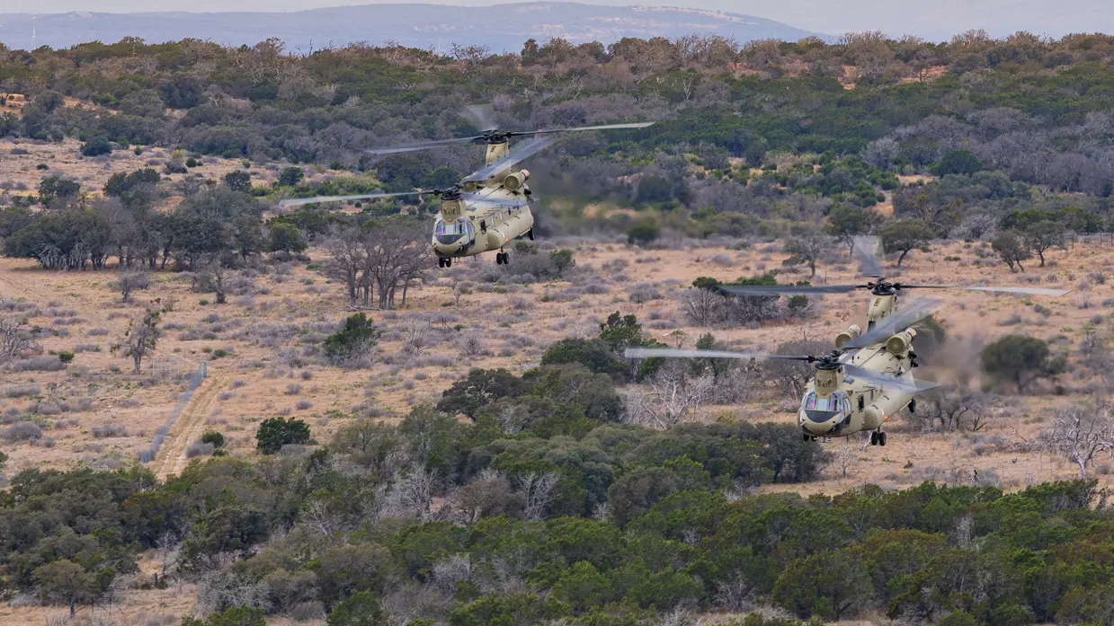 Twee laagvliegende Chinooks boven uitgestrekt landschap met zand en plukken begroeiing. Op de achtergrond heuvels.