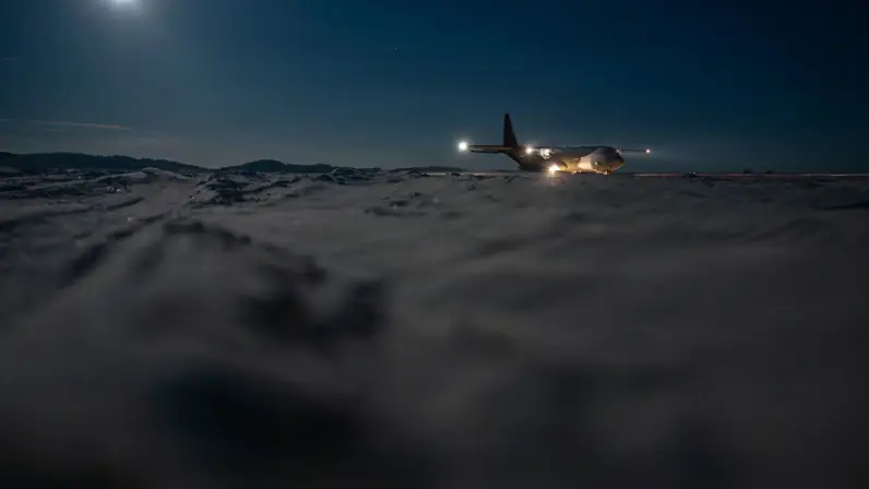 Een militair transportvliegtuig op besneeuwde baan ’s in de nacht, verlicht door eigen lichten, met volle maan en donkere bergruggen op de achtergrond.