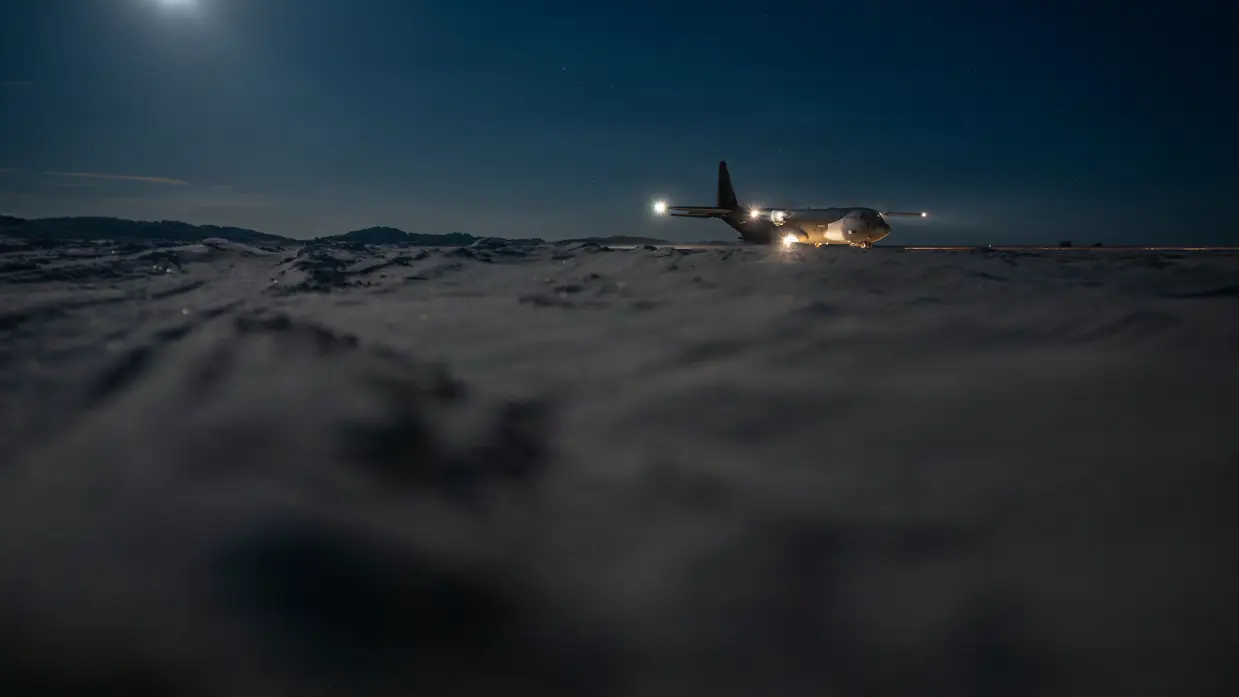 Een militair transportvliegtuig op besneeuwde baan ’s in de nacht, verlicht door eigen lichten, met volle maan en donkere bergruggen op de achtergrond.