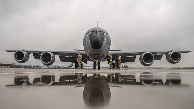 Een groot grijs militair vliegtuig met vier motoren staat op natte startbaan. Drie militairen in camouflage staan erbij.