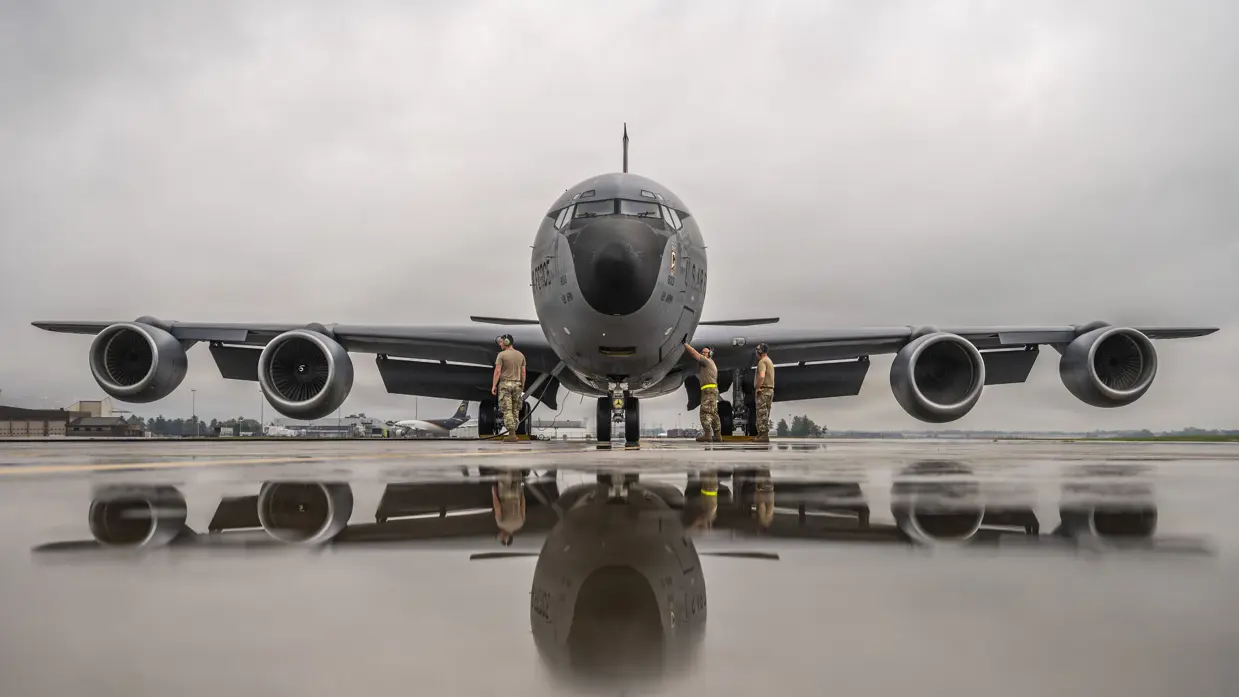 Een groot grijs militair vliegtuig met vier motoren staat op natte startbaan. Drie militairen in camouflage staan erbij.