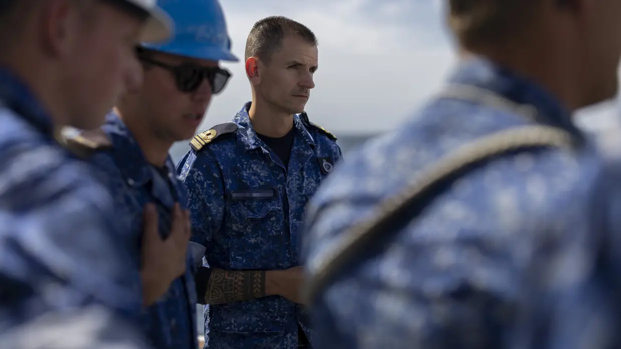 Een officier van de marine in blauw uniform. Hij staat op het dek van een schip en is omringd door matrozen. Hij kijkt strak vooruit.