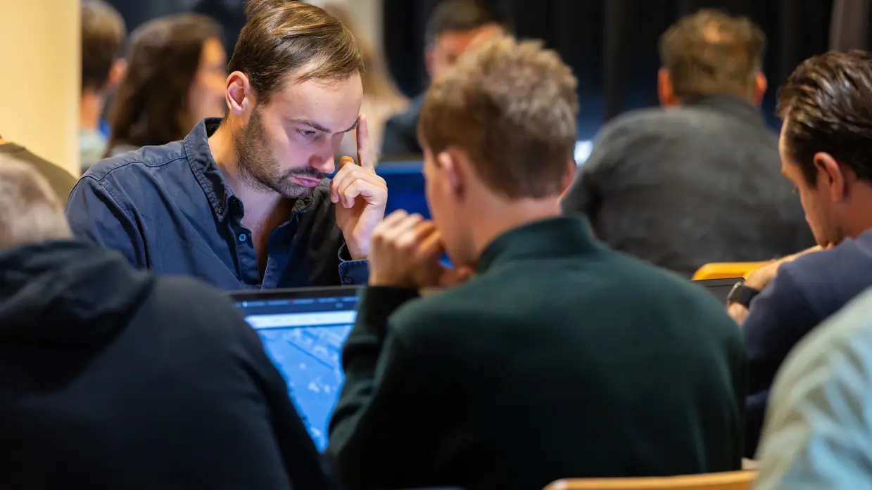 Twee mannen zitten tegenover elkaar en kijken naar hun laptopscherm. Ze zijn omringd door anderen in dezelfde houding.