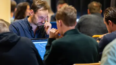 Twee mannen zitten tegenover elkaar en kijken naar hun laptopscherm. Ze zijn omringd door anderen in dezelfde houding.