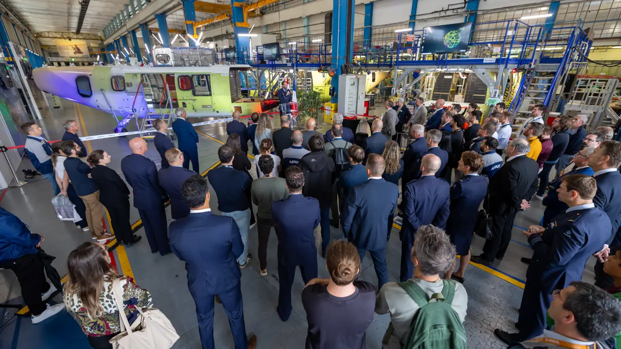 Een man in luchtmachtuniform spreekt een groep aanwezigen toe door een microfoon. Achter hem het frame van de nieuwe helikopter.