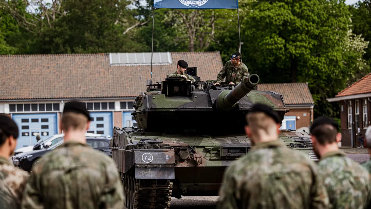 Een Leopard 2A8-gevechtstank met daarop 2 militairen tijdens een ceremonie. Ervoor staan militairen die het voertuig aanschouwen.