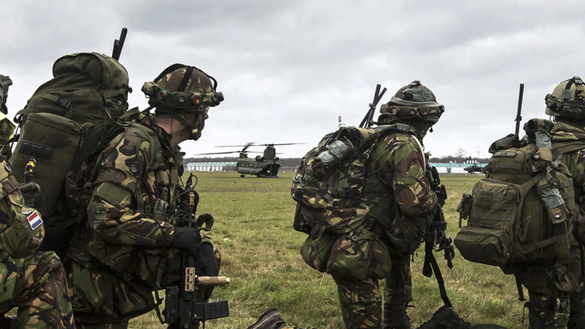 Militairen staan op grasveld en kijken toe hoe een Chinook landt.