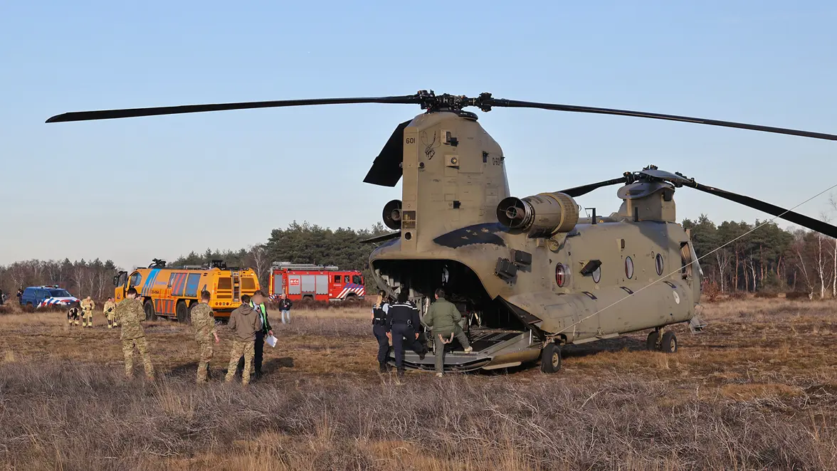 Een militaire helikopter staat in een veld, met daar omheen veel mensen en een gele brandweerauto.