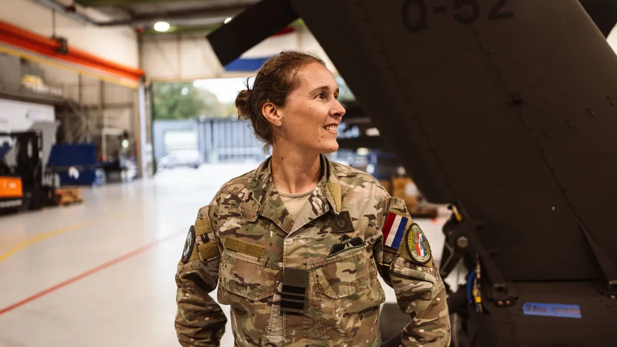 Portretfoto van Majoor Saskia met op de achtergrond het staartstuk van een Apache die in een loods staat.