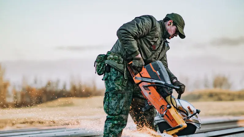 Een militair zaagt met een vonkende zaag een metaalrail buiten in de natuur. 