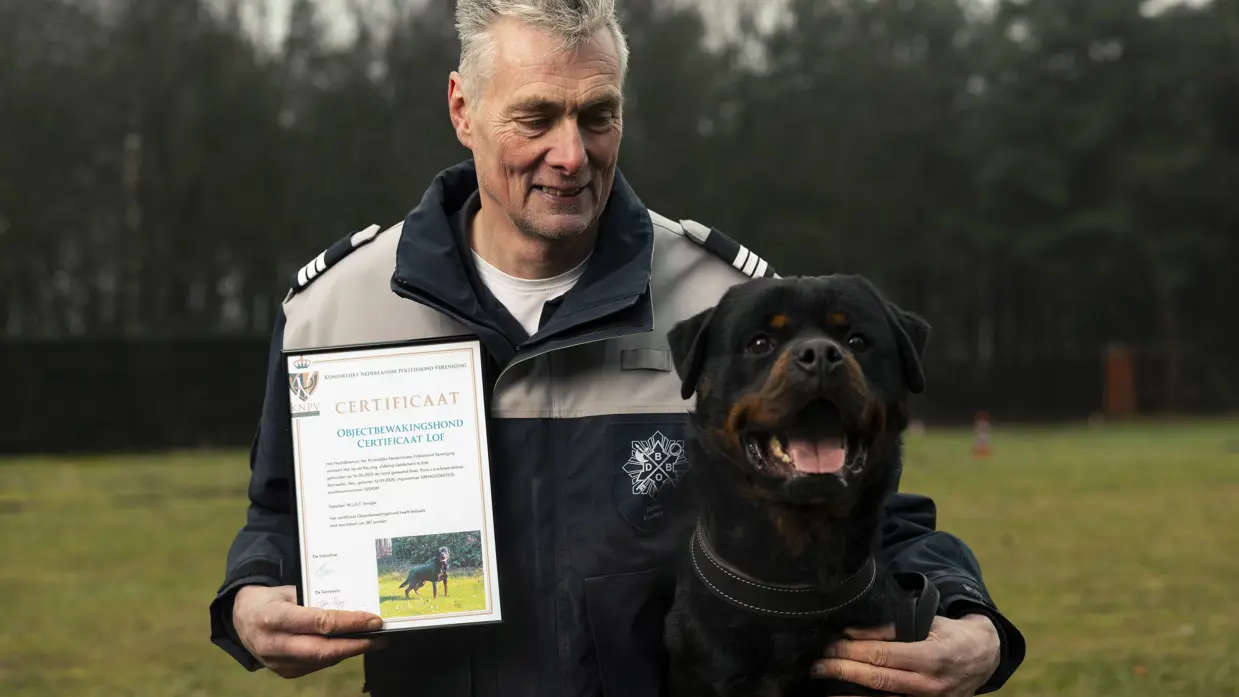 +alttekst: Man, geknield in uniform, houdt trots een certificaat vast. Naast hem zit een rood-zwarte Rottweiler met uitstekende tong in een groene, bosrijke omgeving. 