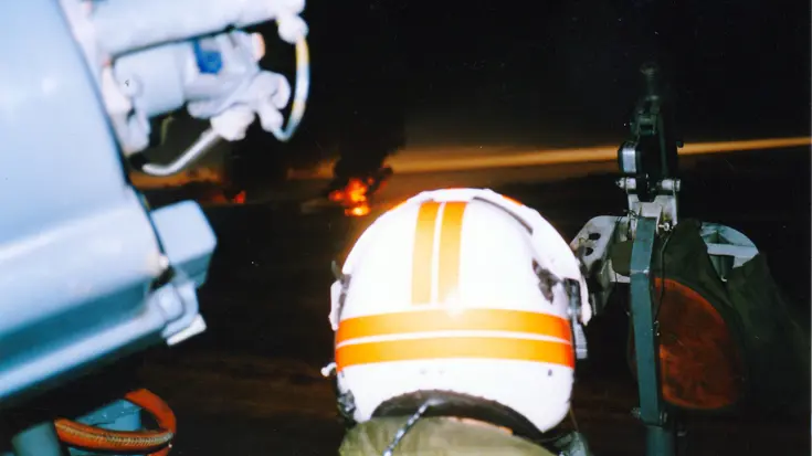 Een bemanningslid van een helikopter, met een witte helm met oranje strepen, kijkt in een donkere lucht met vuur en rook op de achtergrond.
