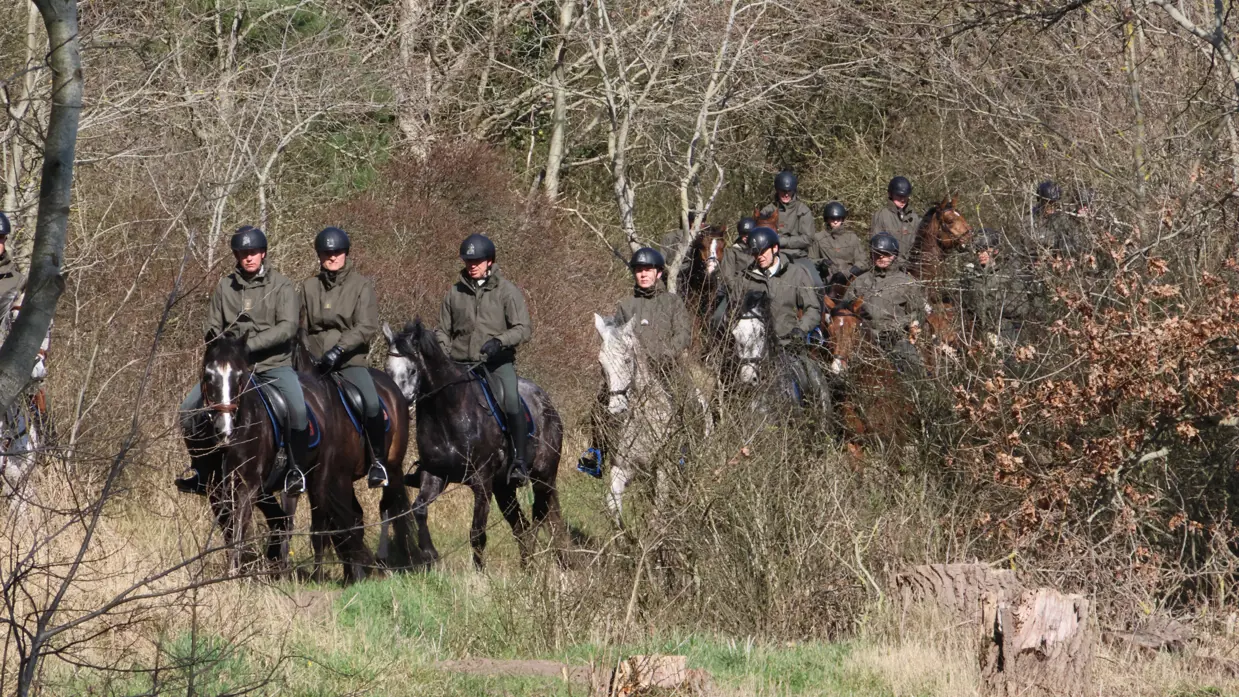 Links: Twee ruiters in uniform en met helmen rijden in galop op respectievelijk een bruin en wit paard over een zandstrand langs de zee. Rechts: Een groep ruiters in militair uniform en met helmen zit te paard in een bosrijk gebied met kale bomen en struiken.