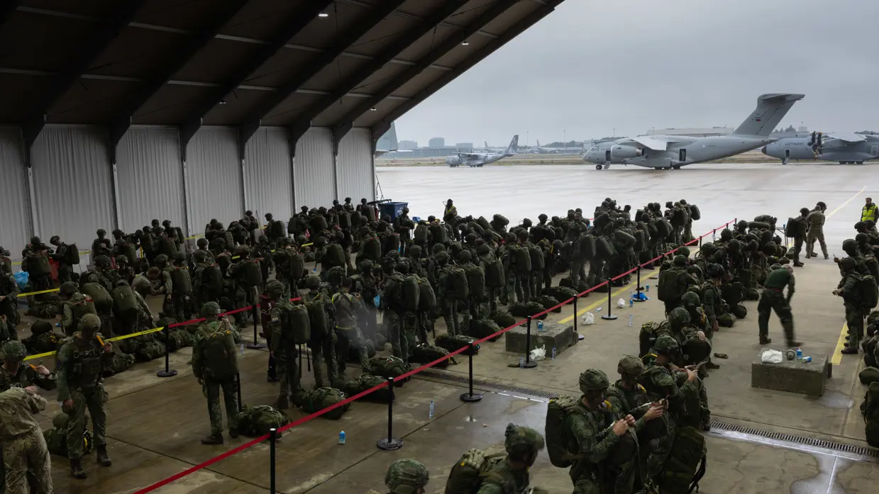 In een grote hangar staan soldaten in camouflagetenue met rugzakken in rijen. Buiten wachten militaire vliegtuigen op het platforms, onder een grijze lucht.