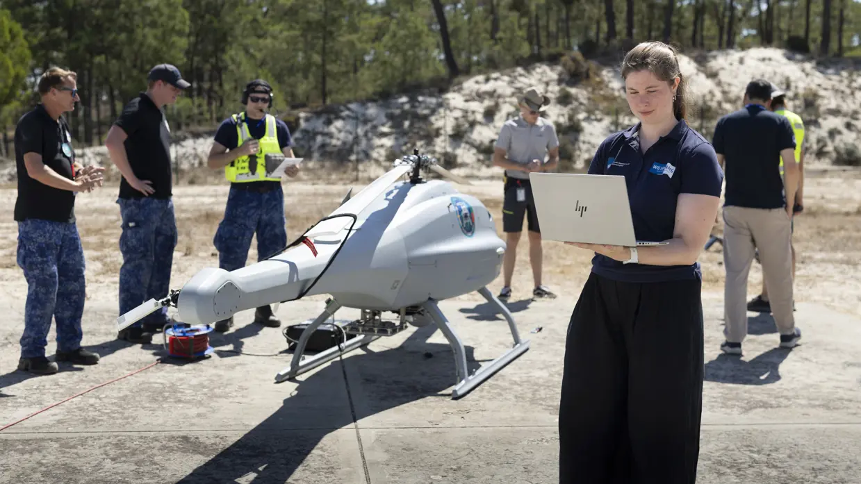 Een vrouw staat met een laptop in haar hand en kijkt naar het scherm. Daarachter een grijze helikopterdrone en mensen in veiligheidshesjes.