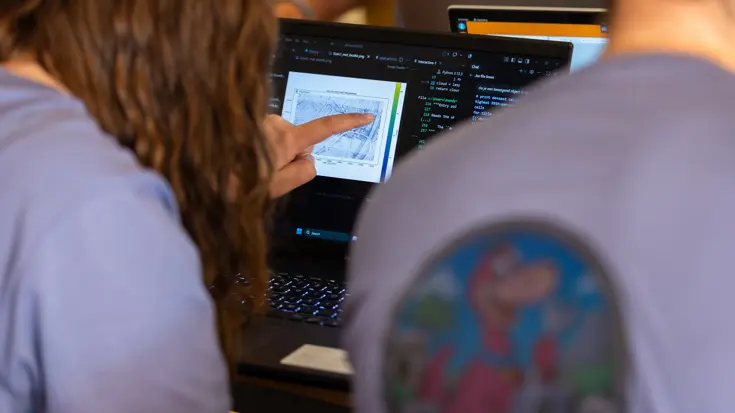 Twee mensen in lilakleurig shirt zitten naast elkaar, terwijl de linker met een vinger wijst op het &nbsp;laptopscherm.