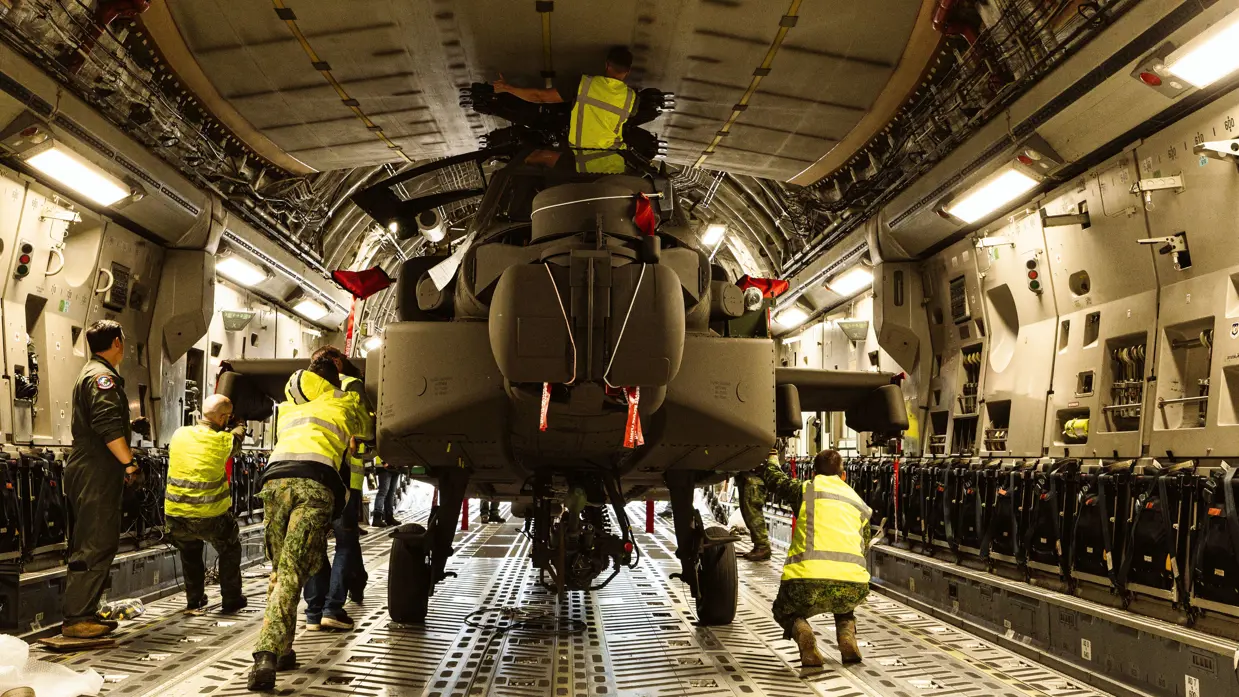 Militairen staan in het laadruim van een vrachtvliegtuig en duwen een Apache naar buiten.