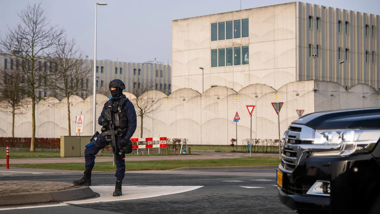 Een zwaarbewapende marechaussee in donkerblauw uniform staat bij een wegversperring naast een groot, grijs gebouw met een zwarte auto op de voorgrond.