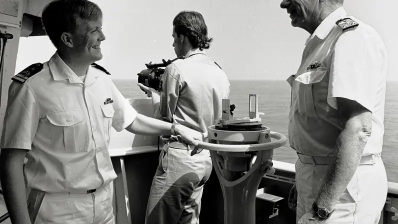 Drie personen in marine-uniform staan op een schip. Twee lachen naar elkaar, één houdt een verrekijker vast. Ze staan bij navigatieapparatuur met zee op de achtergrond.