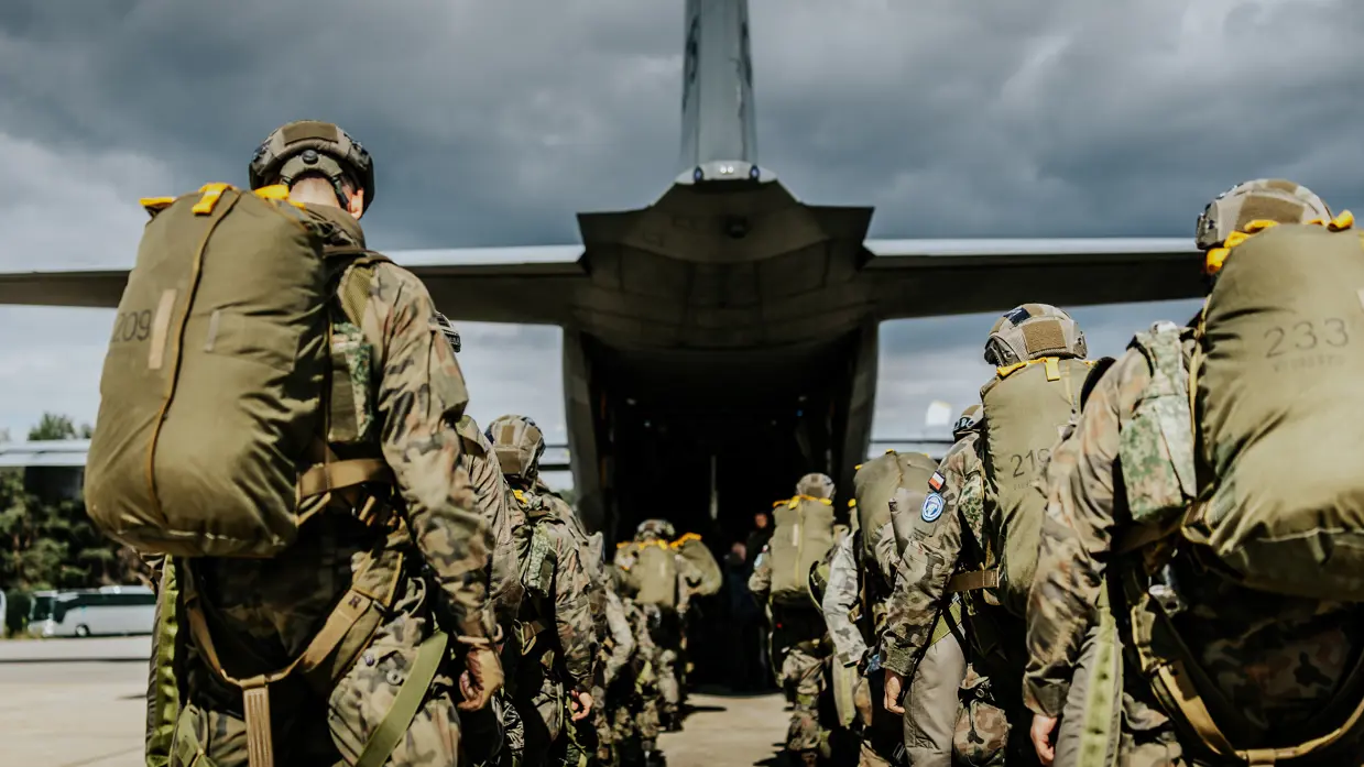 Soldaten met camouflagetenue, helmen en grote rugzakken lopen in een rij over een vliegveld naar een groot grijs militair vliegtuig met open laadklep, onder bewolkte lucht.