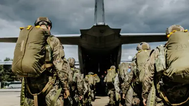 Soldaten met camouflagetenue, helmen en grote rugzakken lopen in een rij over een vliegveld naar een groot grijs militair vliegtuig met open laadklep, onder bewolkte lucht.