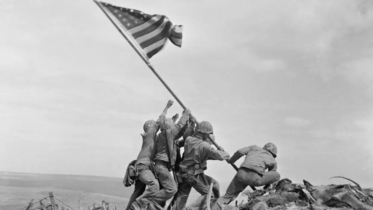 Zes Amerikaanse soldaten werken samen om een grote Amerikaanse vlag op een metalen paal te hijsen, hoog op een rotsachtige heuvel. De wind laat de vlag wapperen. Rechts het monument. 