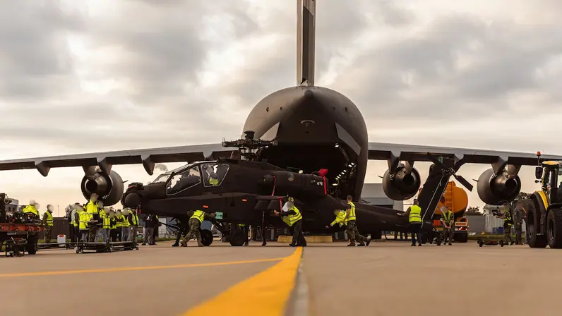 Militairen in gele hesje duwen een Apache voort, op de achtergrond gaapt het open laadruim van het C17 transportvliegtuig.