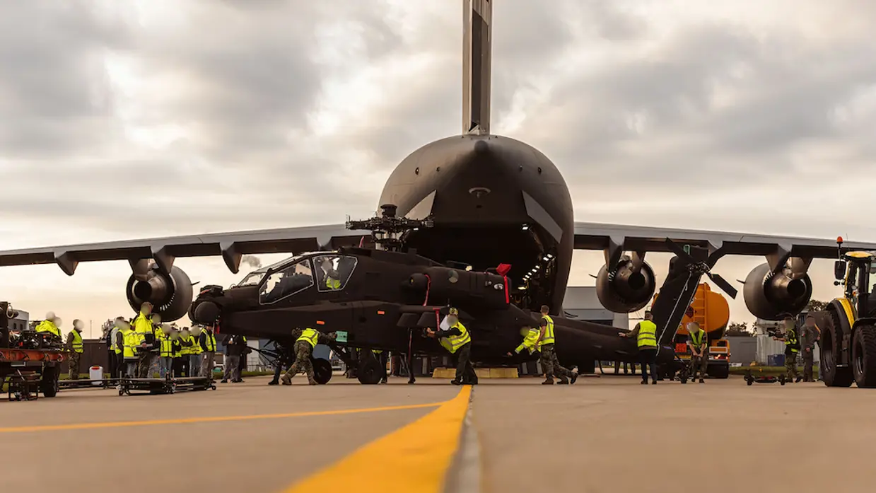 Militairen in gele hesje duwen een Apache voort, op de achtergrond gaapt het open laadruim van het C17 transportvliegtuig.