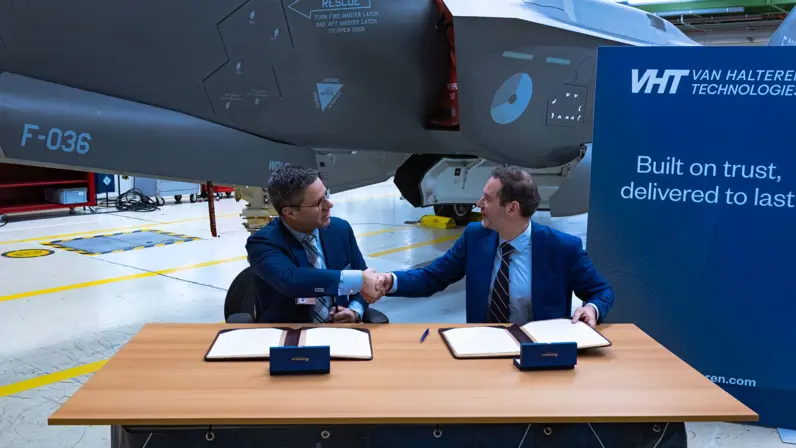 Twee mannen in pak schudden elkaar de hand. Op de tafel waaraan zij zitten liggen contracten. Op de achtergrond staat de replica van de F-35.