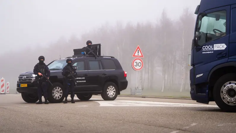 Drie zwaarbewapende agenten in donker uniform staan bij een zwarte politieauto; een vrachtwagen rijdt voorbij op een mistige weg met maximumsnelheid 30.