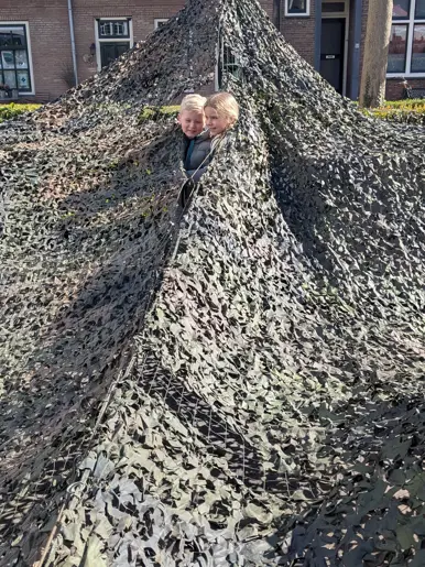 Links: Twee glimlachende kinderen kijken onder een grote, groene camouflagenetten-tent vandaan, die buiten voor huizen is opgebouwd en lijkt op een militaire schuilplaats. Rechts: Een glimlachend kind in een militair kogelwerend vest en helm geeft een volwassene in uniform een high-five op een buitenlocatie.