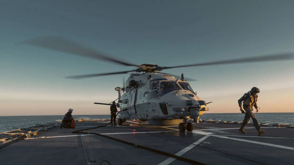 Een grijze militaire helikopter landt op het dek van een schip terwijl twee mannen met helmen op wegrennen.