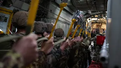 In een militair vliegtuig staan soldaten in camouflagetenue en helmen in een rij, vasthoudend aan gele hangriemen vanaf het plafond.