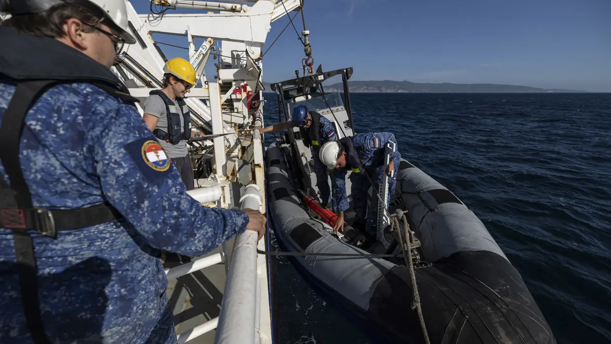 Een RHIB-motortboot hangt aan de zijkant van een schip. Op het bootje staan twee matrozen van de marine. Een vrouw in grijs t-shirt en man in blauw uniform kijken toe. 