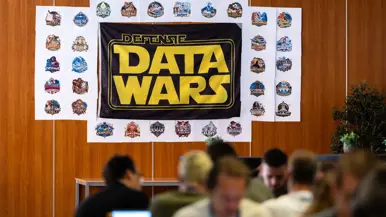 Een vlag van de Defensie Data Wars hangt aan een houten muur met op de voorgrond mensen achter een laptop.