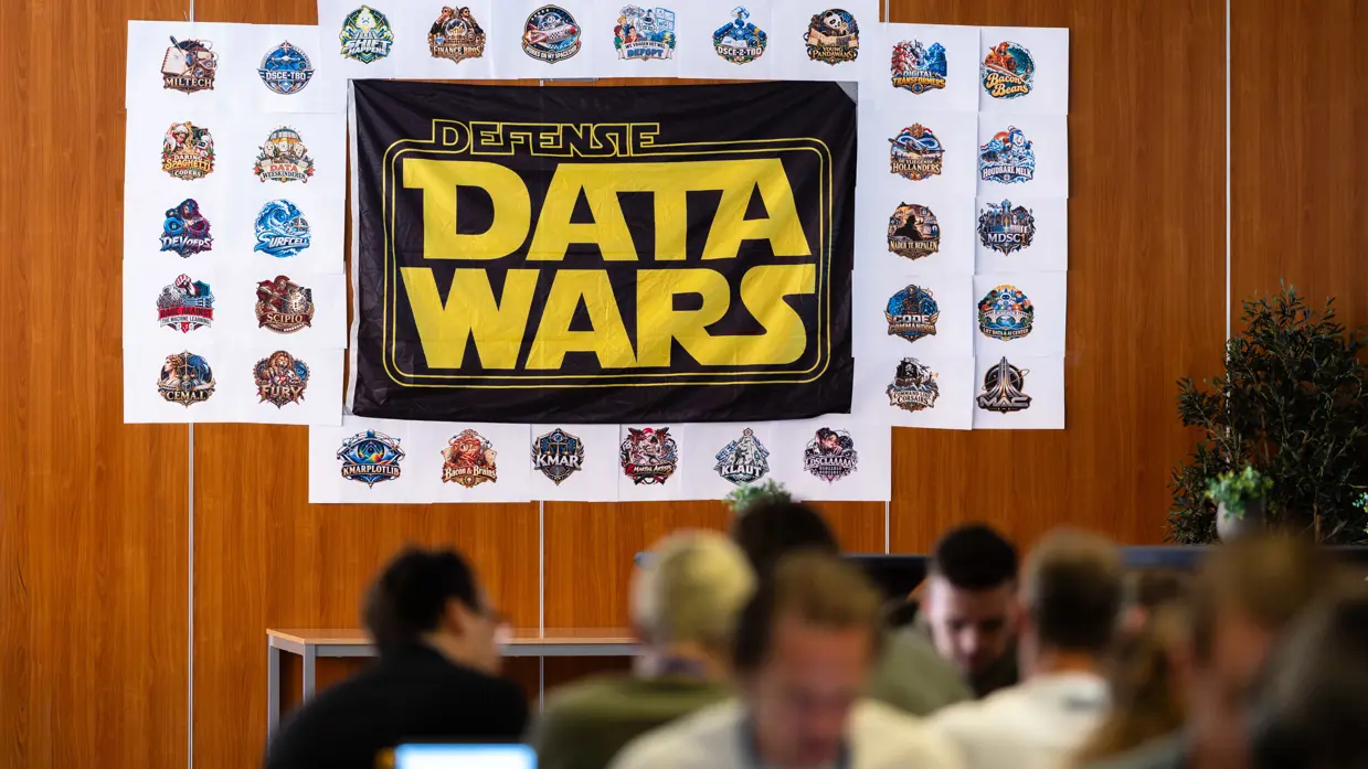 Een vlag van de Defensie Data Wars hangt aan een houten muur met op de voorgrond mensen achter een laptop.