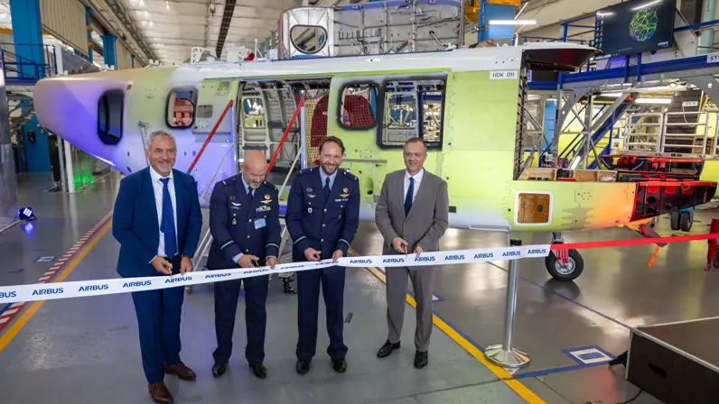 Vier mannen in pak, van wie twee militairen, knippen een lintje door. Op de achtergrond het helikopterframe.