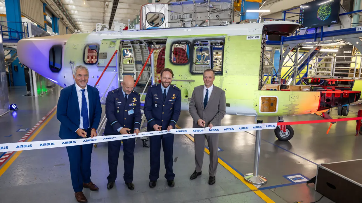 Vier mannen in pak, van wie twee militairen, knippen een lintje door. Op de achtergrond het helikopterframe.