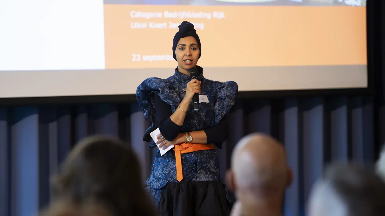 Een vrouw met een blauwe tulband en een blauwe jurk spreekt met een microfoon tijdens een presentatie. Op de achtergrond is een groot scherm zichtbaar.